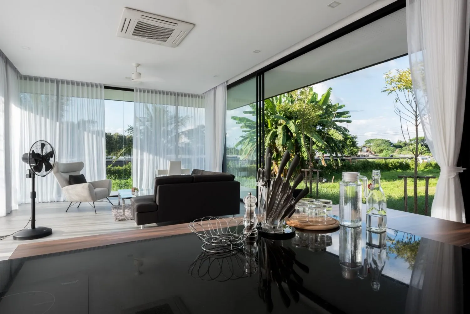 Modern living room with glass walls and furniture, large windows overlooking greenery.