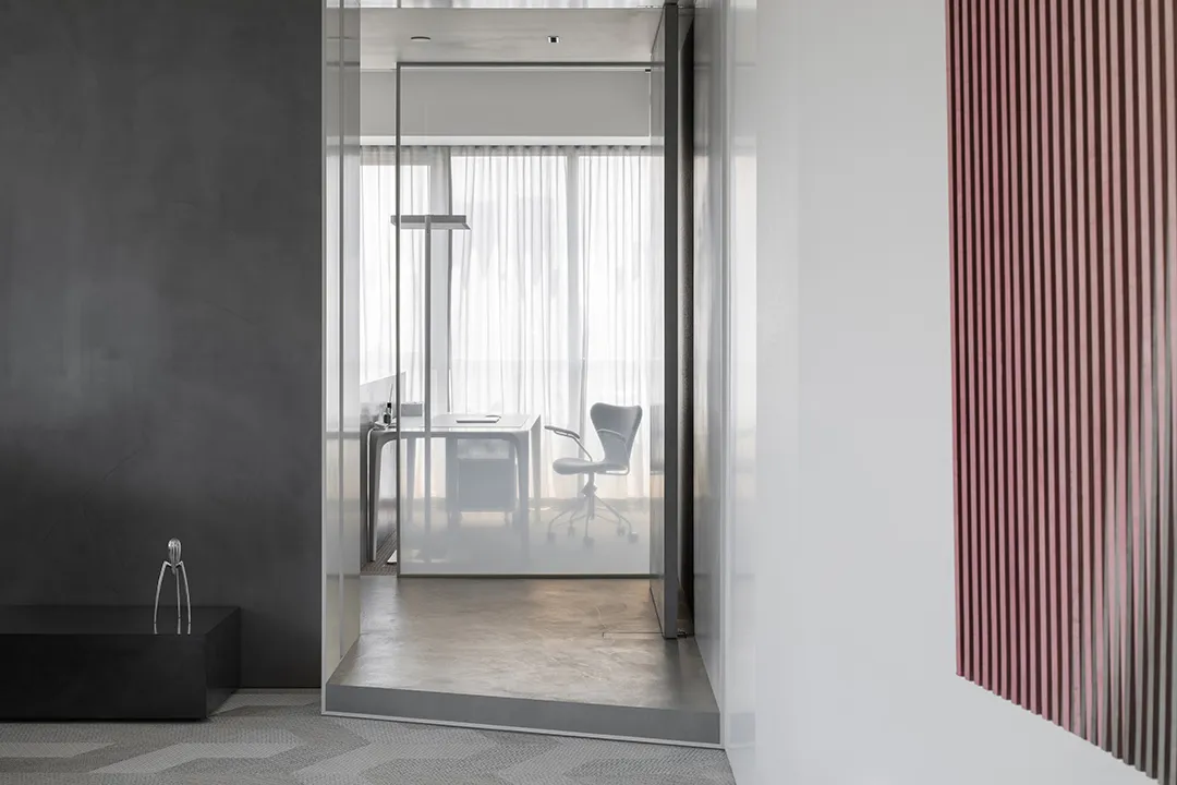 A minimalist office interior featuring glass walls and concrete flooring, with a single chair and desk.