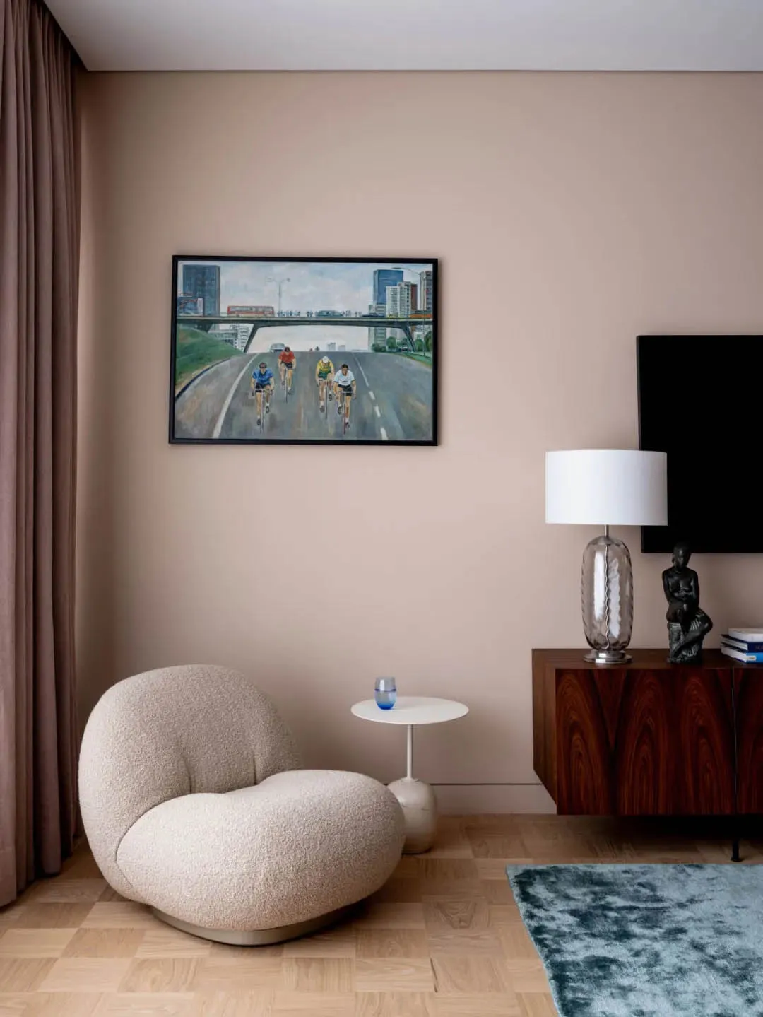 A modern living room with a sand-colored plush chair, a white circular side table, a wooden sideboard with a lamp, and a larg