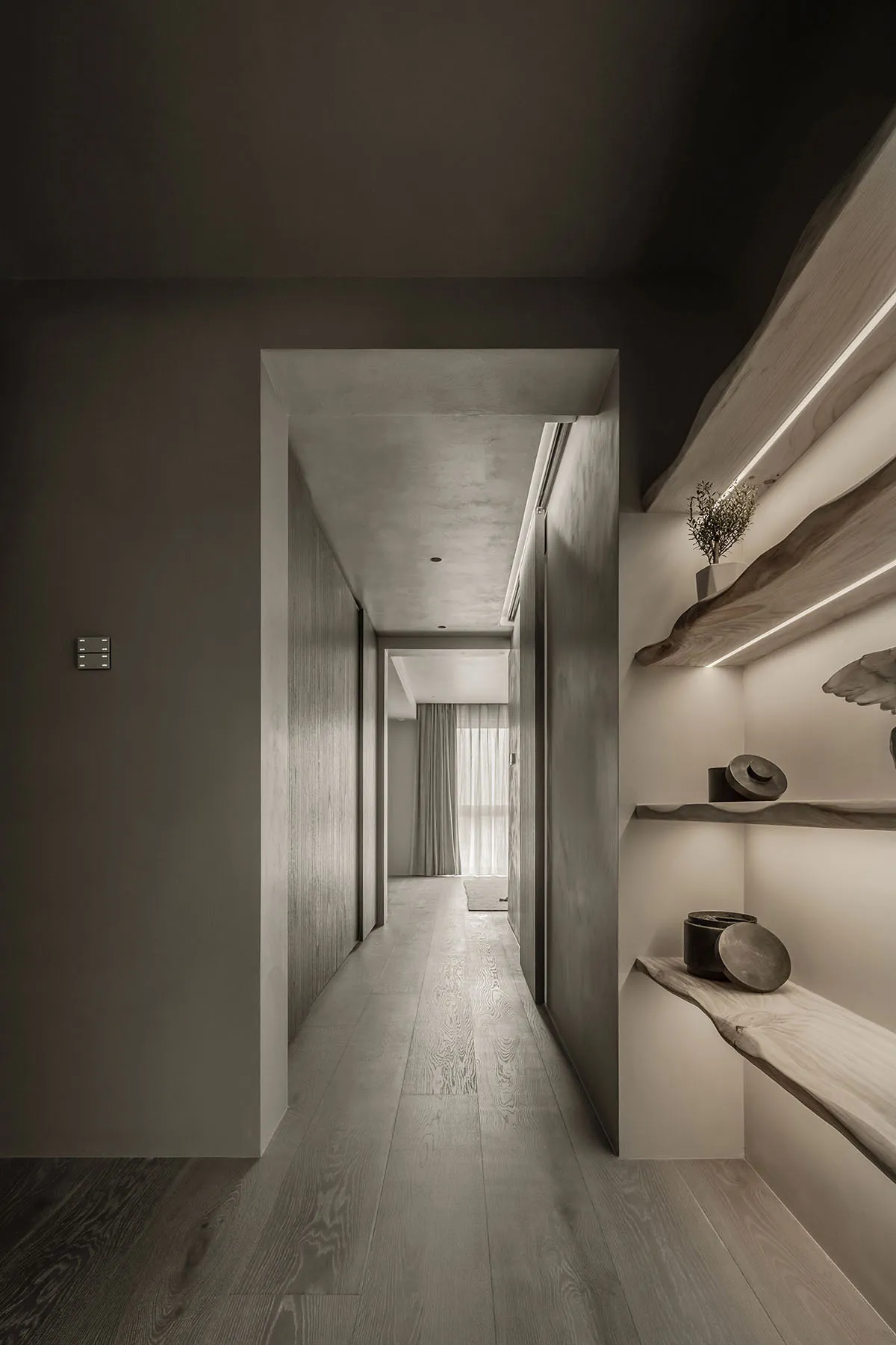 A modern corridor featuring light wood flooring and walls, with a subtle decorative shelf and natural lighting from a window