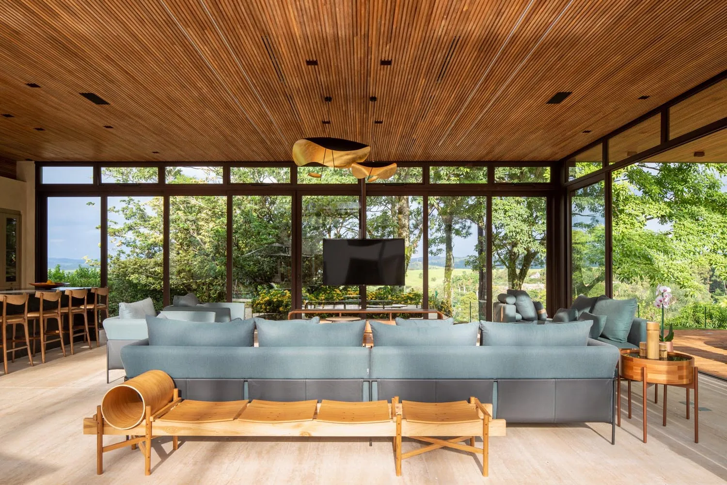Modern living room featuring a blue sofa, wooden chairs, and a TV mounted above a table, with large windows providing a sceni