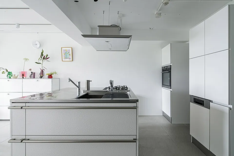 A modern, minimalist white kitchen with a cooking area in the center, featuring a double-sided stainless steel countertop, an