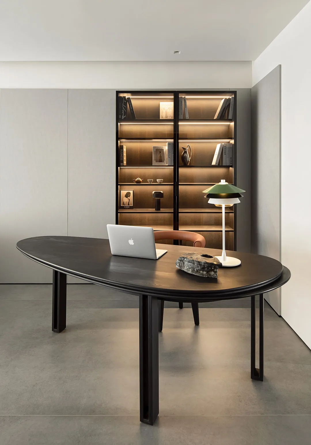 A minimalist office setting featuring a wooden desk with a laptop, lamp, and decorative rock. Behind the desk is a well-lit b