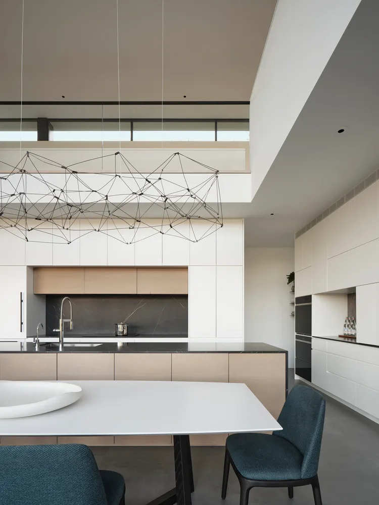 A modern minimalist kitchen with white cabinets, a white table, and blue fabric chairs, featuring a geometric chandelier and