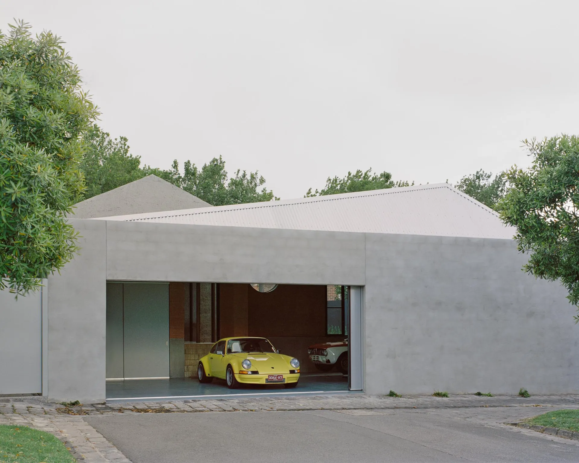 A modern concrete garage with a yellow car parked inside, surrounded by greenery and a paved driveway.