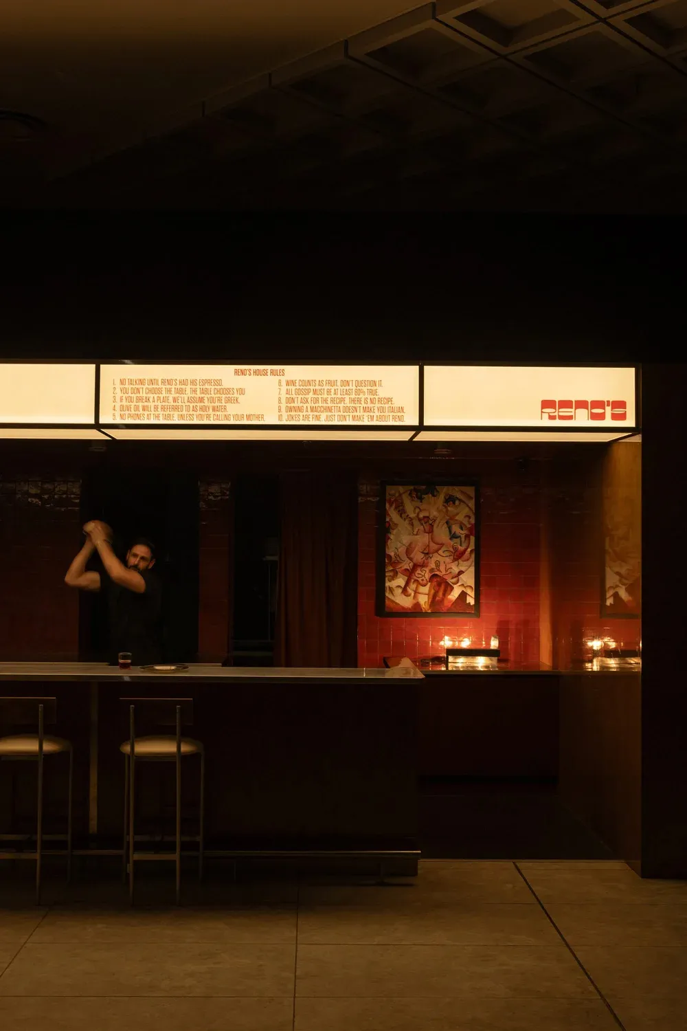 A casual bar with a brick wall, two chairs, and a counter with wine glasses, a cup, and a bowl, with a man standing behind the counter