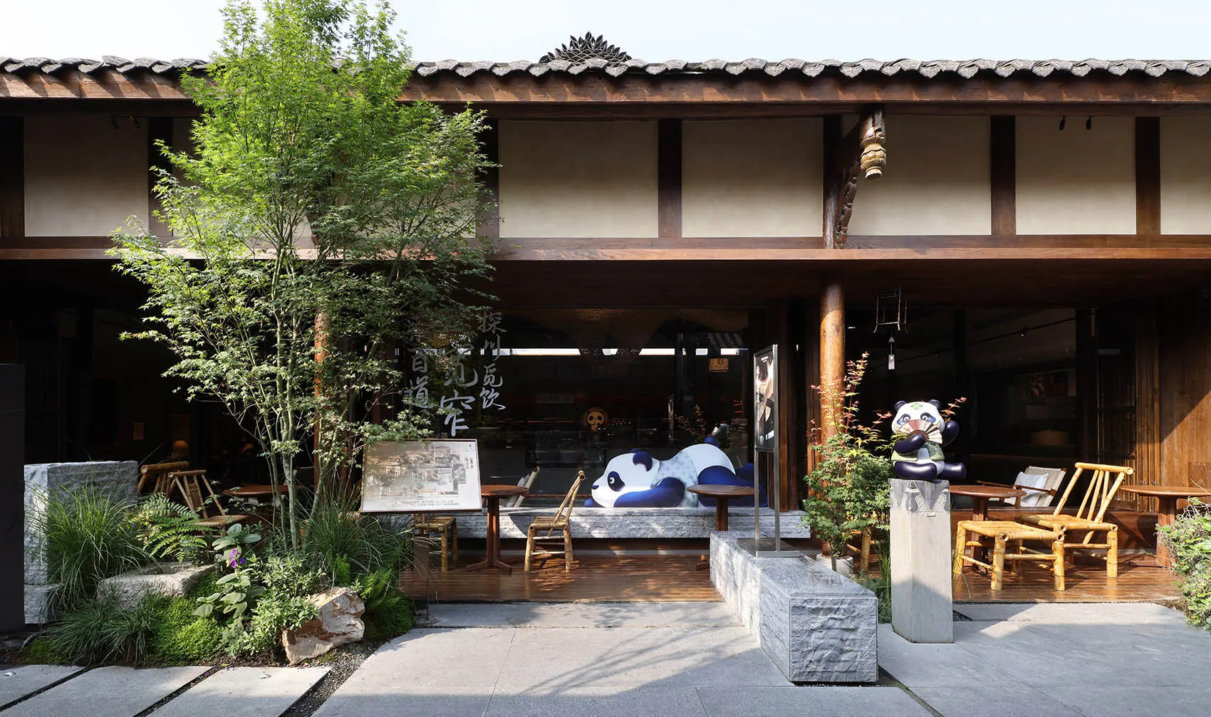 An inviting outdoor seating area with a wooden building facade, a stone table, and a large panda bear sculpture, surrounded by chairs and Asian design elements