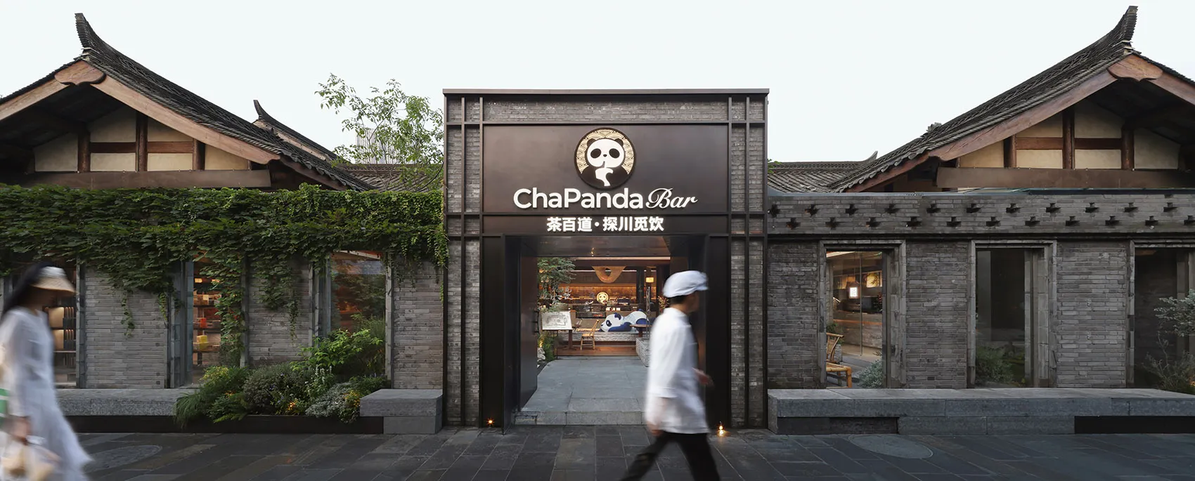 A man walks in front of an Asian restaurant with a black facade, featuring dining tables and chairs inside, creating an inviting and comfortable atmosphere