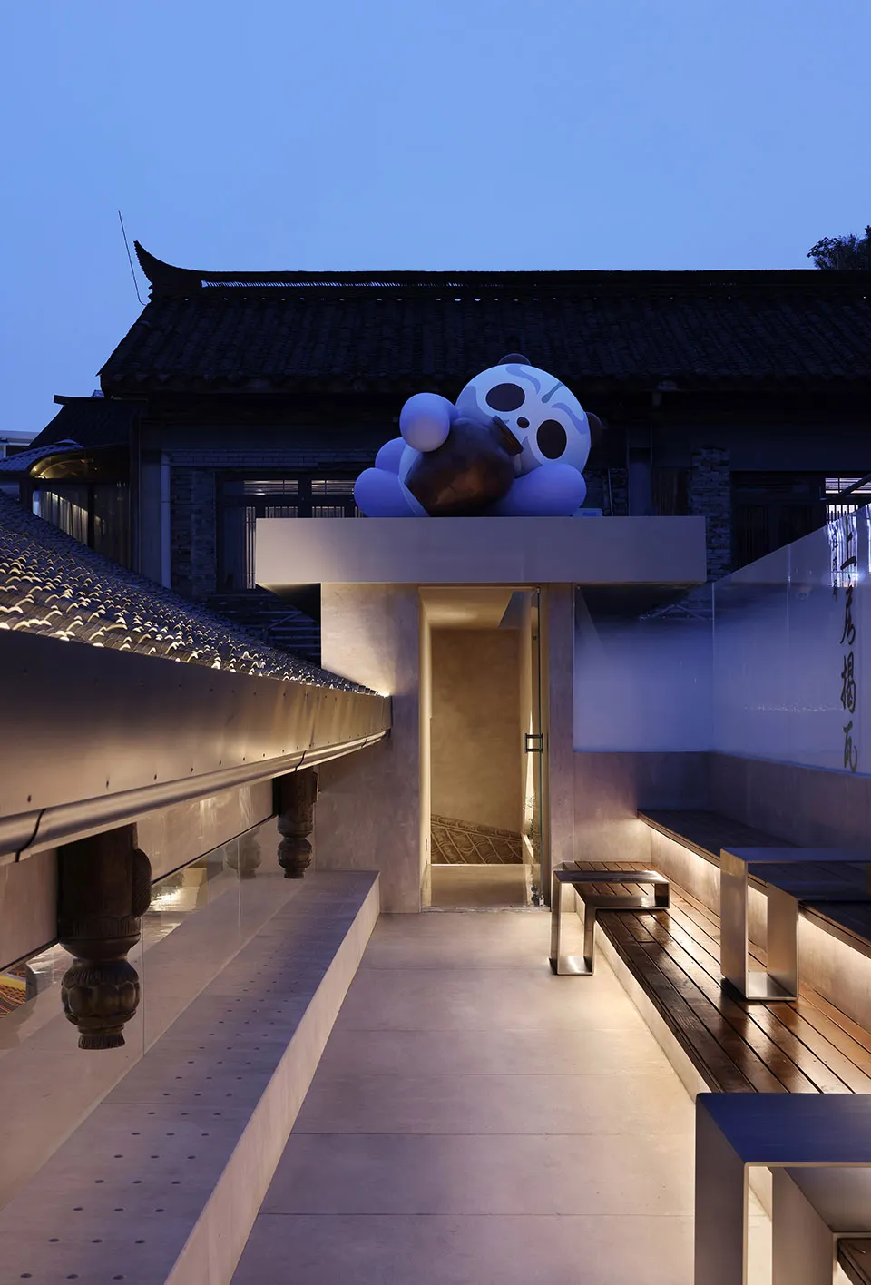 A serene Asian-style building with a stone panda statue on a balcony, overlooking a bridge and street below, surrounded by benches in a peaceful environment