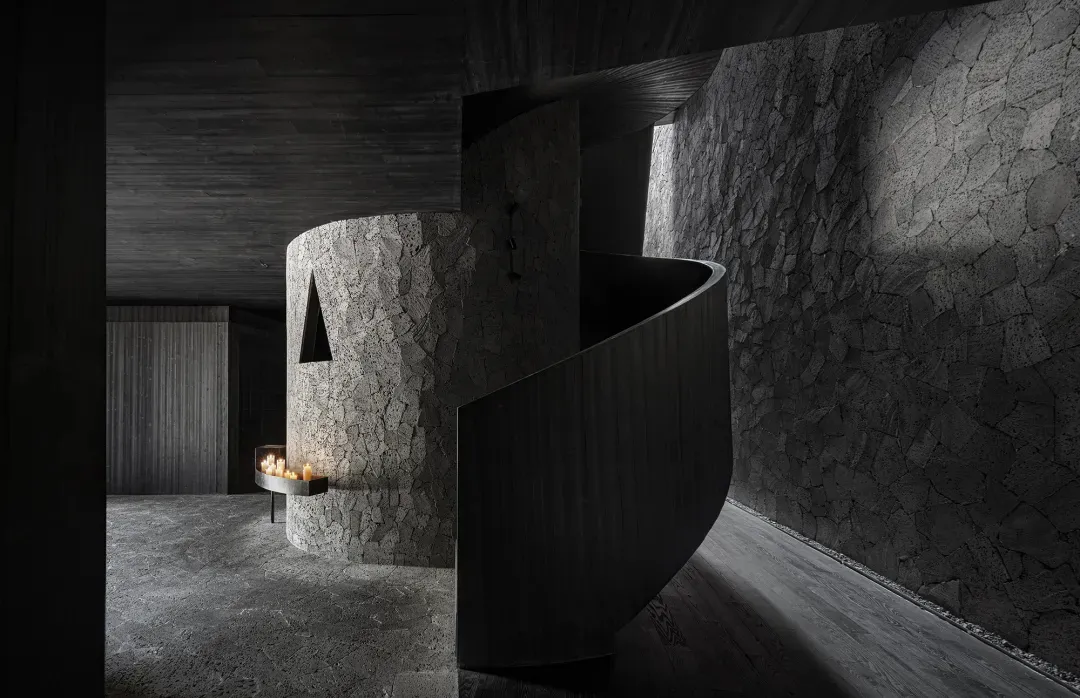A modern living space with a stone spiral staircase, cave-like walls, a fireplace, and minimalist decor