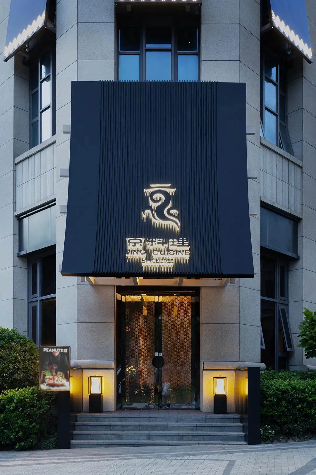 Exterior view of an Asian restaurant with a stone facade, black awning, and potted plants, featuring a clock on the wall and an inviting atmosphere