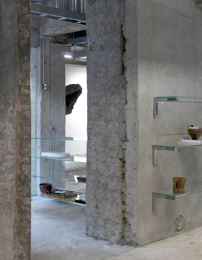 A well-lit art gallery with a display case filled with vases, bowls, and books, set against industrial concrete walls and illuminated by overhead lights