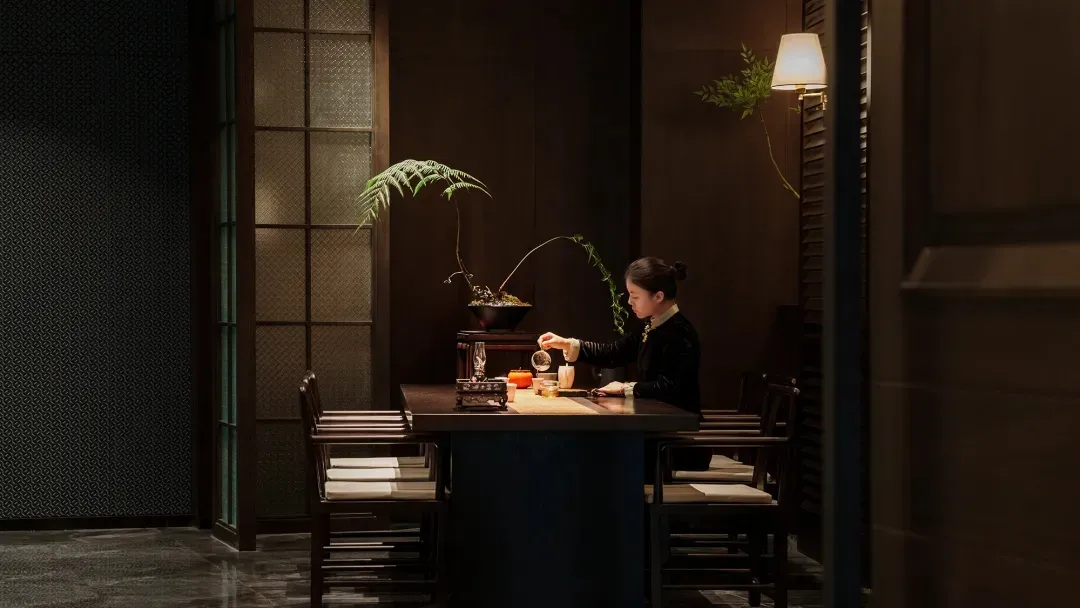 一位女性坐在餐桌旁，置身於燈光昏暗的房間，周圍環繞著椅子，營造出溫馨親密的氛圍