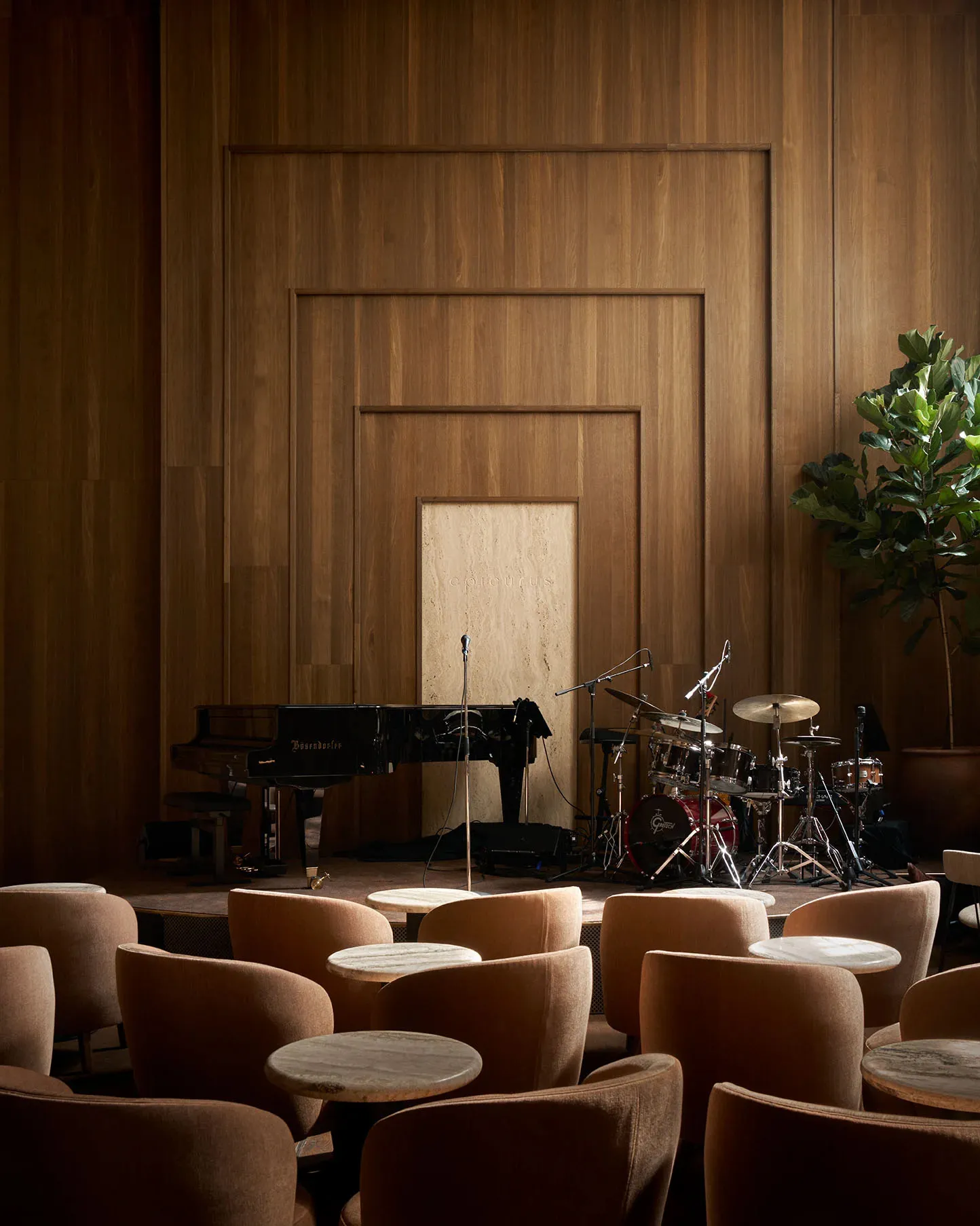 A modern room with wooden walls and floors, featuring a piano and drum set, chairs, and potted plants, creating a well-lit and comfortable atmosphere for musical performances or gatherings