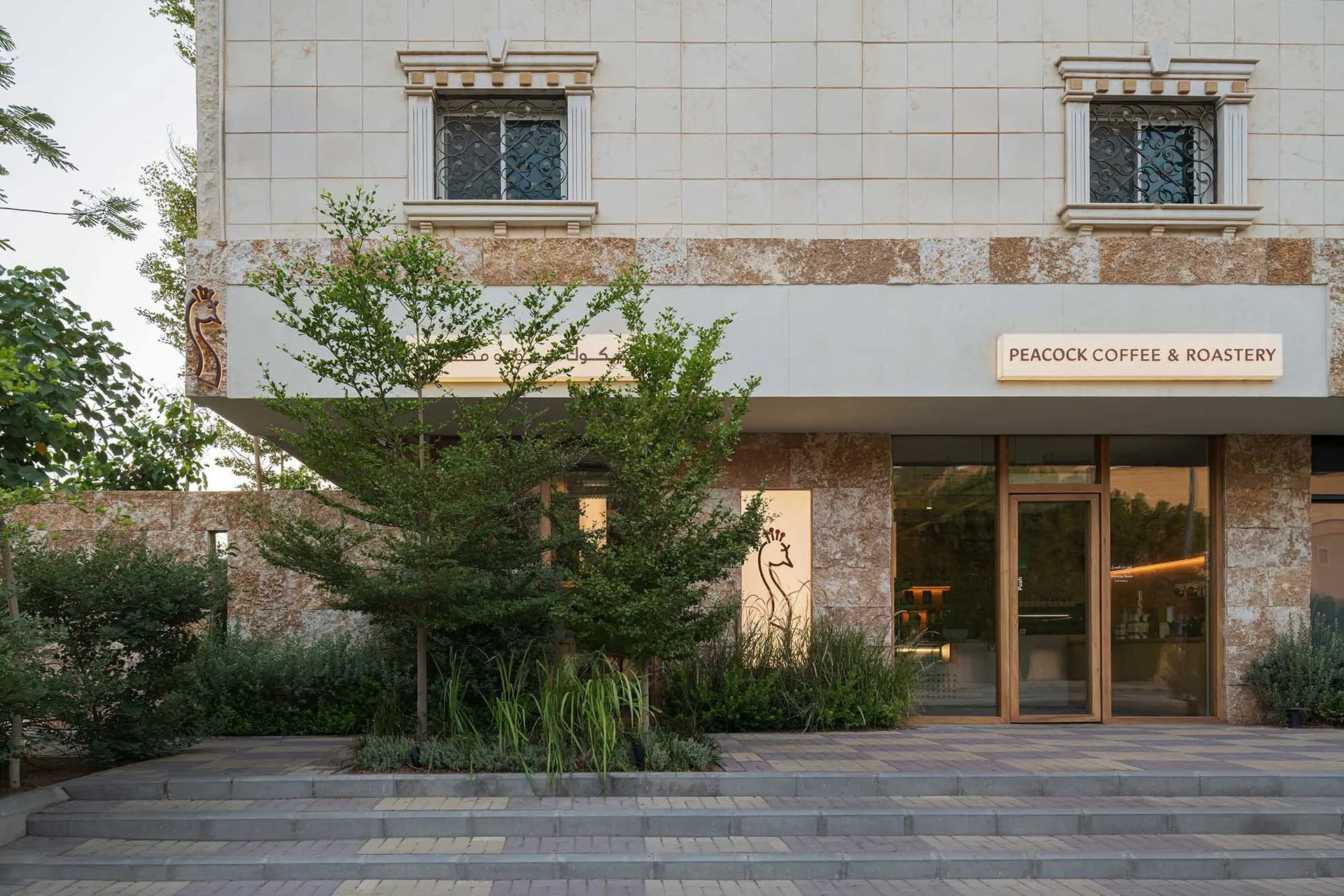 A stone building facade with a large window, surrounded by two trees in a serene environment