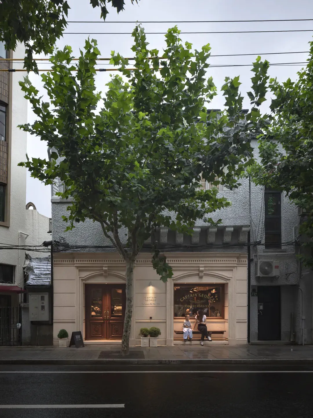 A serene outdoor scene of a building with a tree in front, featuring a white facade, two people, and a bench, set against a calm atmosphere