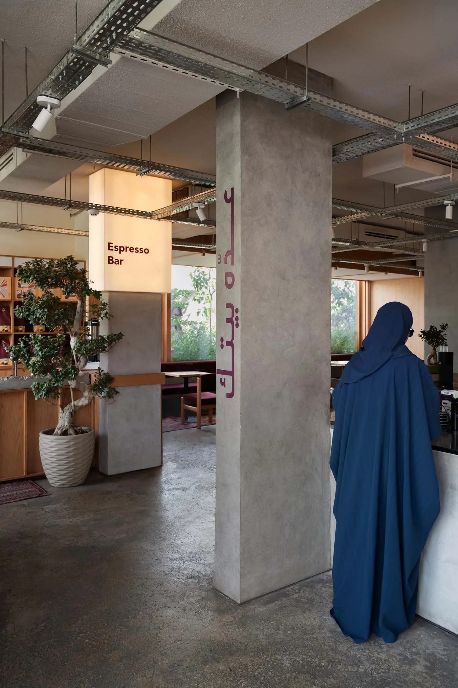 A modern cafe interior with concrete walls, wood accents, and metal elements, featuring a woman standing at a counter and several chairs for seating, with a potted plant in the background