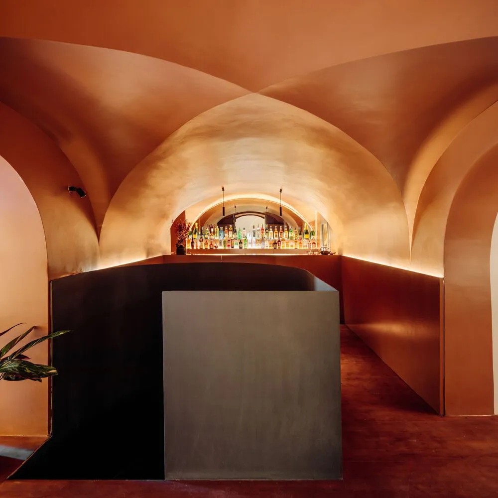 Dimly lit bar area with a long counter, bottles, and wine glasses, surrounded by red-hued walls