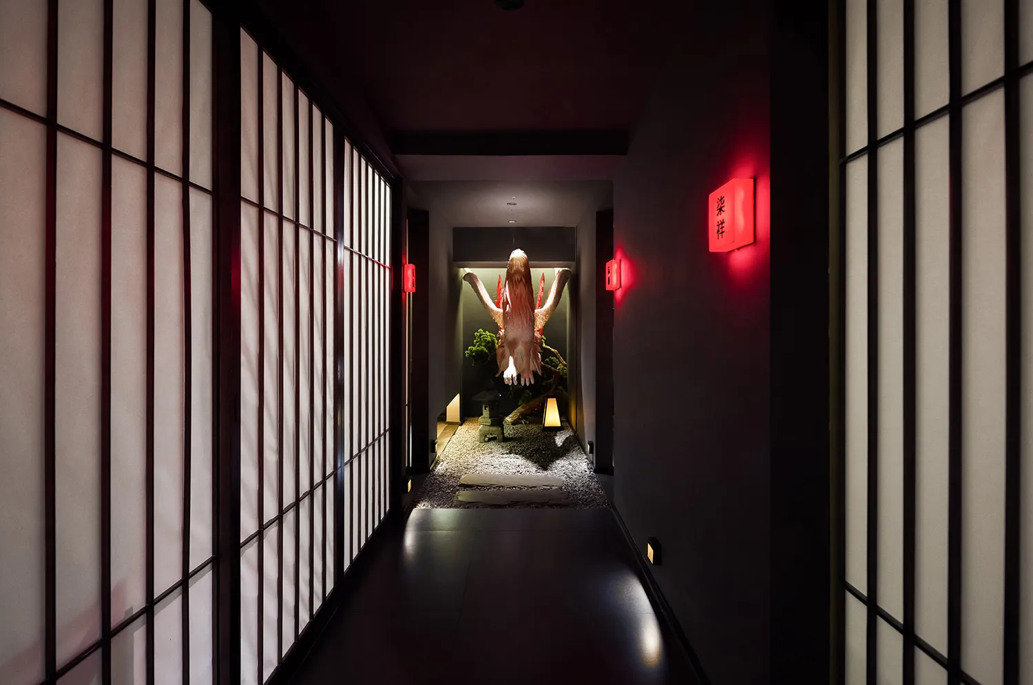 A dimly lit hallway in an Asian home features a woman in traditional clothing standing near a doorway, surrounded by a red accent wall, potted plants, and a decorative vase