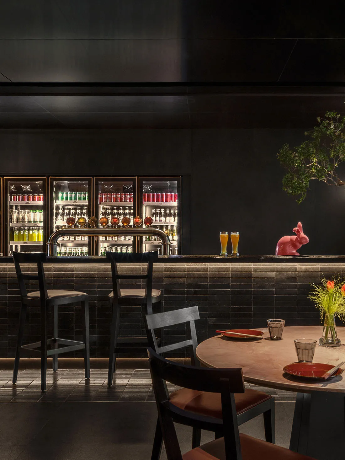 A cozy bar area with a counter, potted plant, and comfortable seating for patrons, surrounded by bottles and an inviting atmosphere
