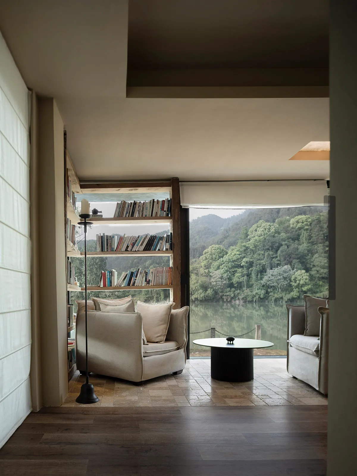 A bright and inviting living space with a large window overlooking a lake, featuring two couches, a chair, and a coffee table, surrounded by books and filled with natural light