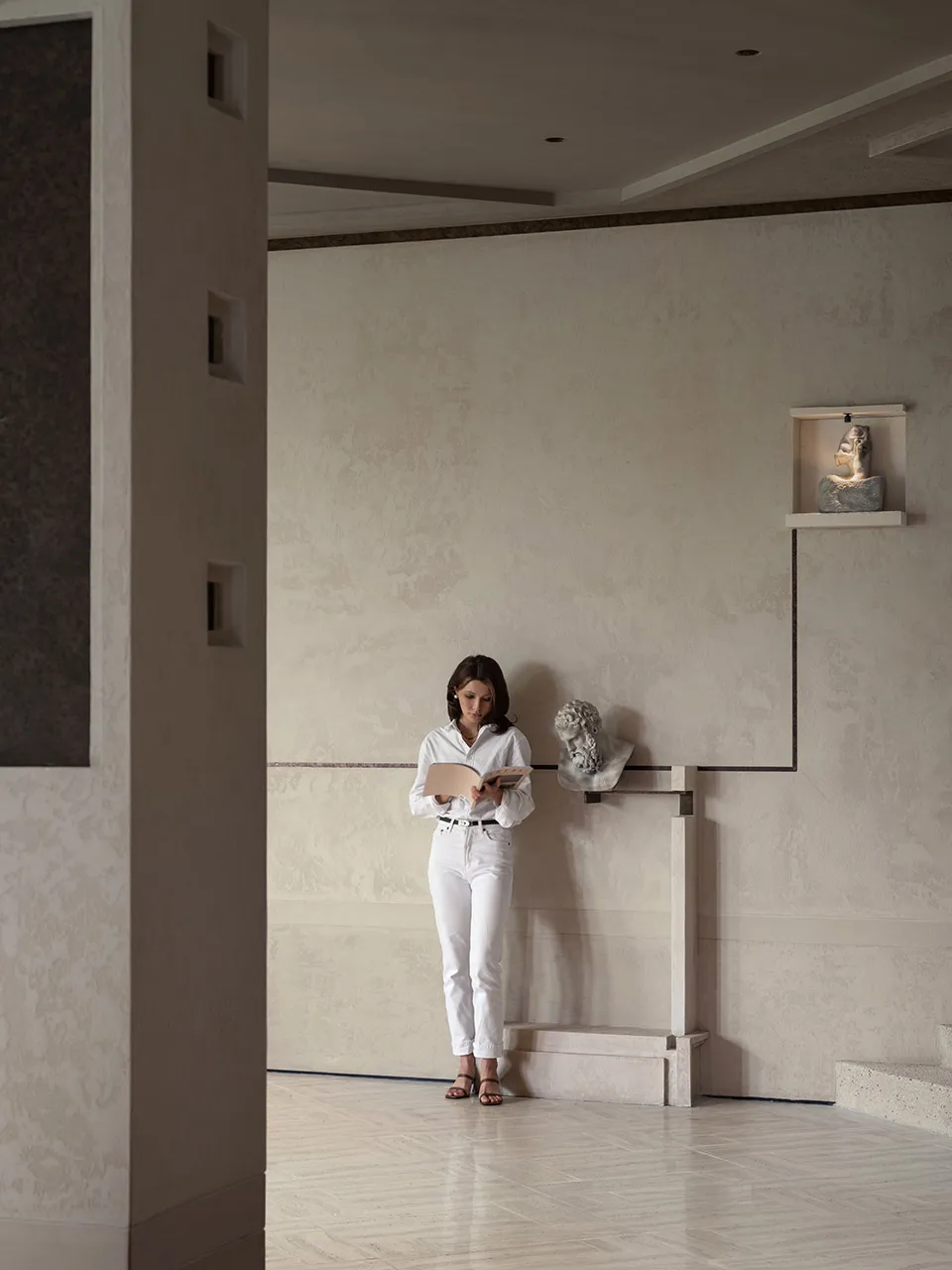 A woman reads a book in a calm, empty room with a white color scheme
