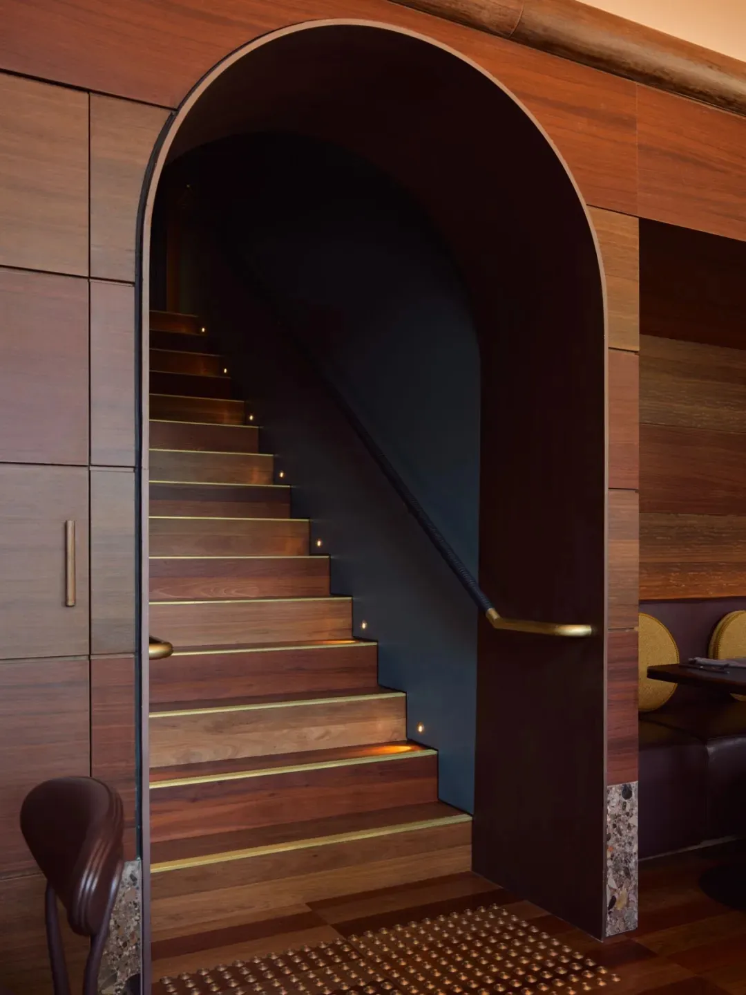 A dark interior staircase with a wooden structure and black railing, flanked by two chairs near the bottom, creating a mysterious atmosphere