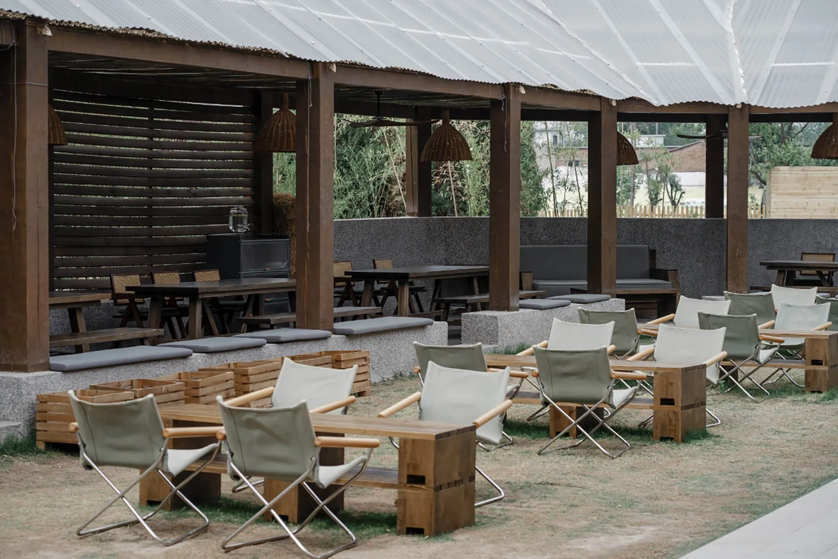 A restaurant patio area with wooden tables and chairs under a large umbrella, set on a dirt ground with a garden-like atmosphere