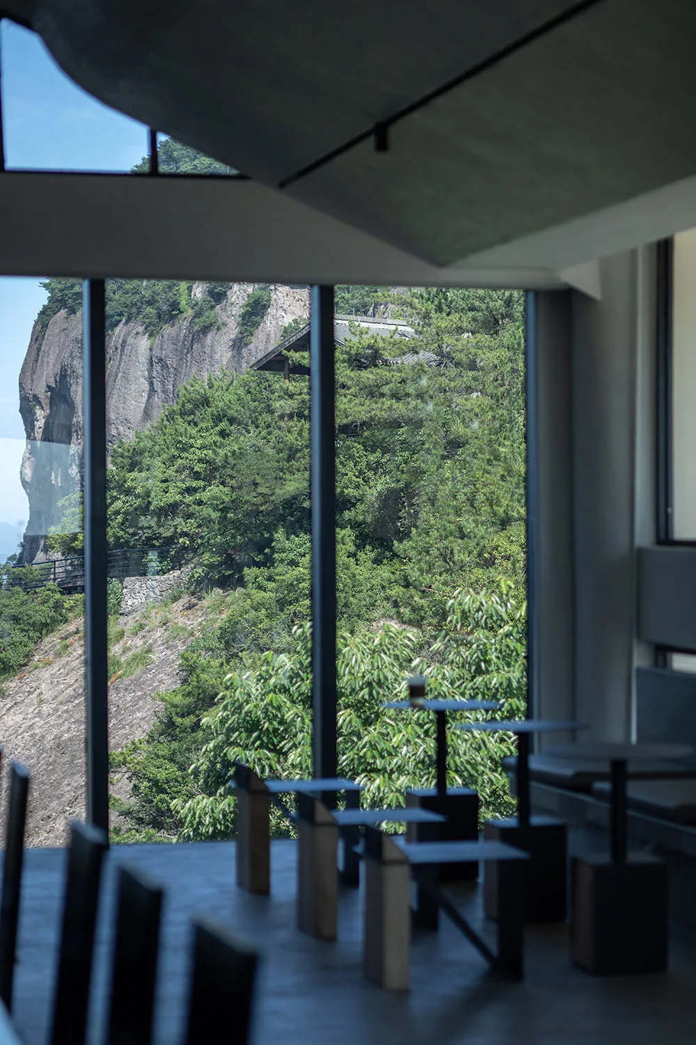 A modern restaurant with glass walls and wooden elements, offering a view of mountains in the distance through large windows