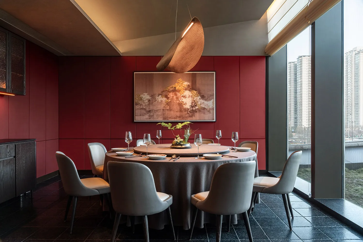 A dining area with a round table set with wine glasses, bowls, and vases, surrounded by chairs, in a room with red walls and potted plants