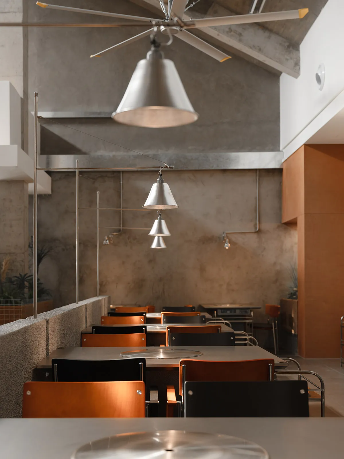 Empty restaurant interior with multiple dining tables and chairs, concrete and wood walls, and overhead lighting