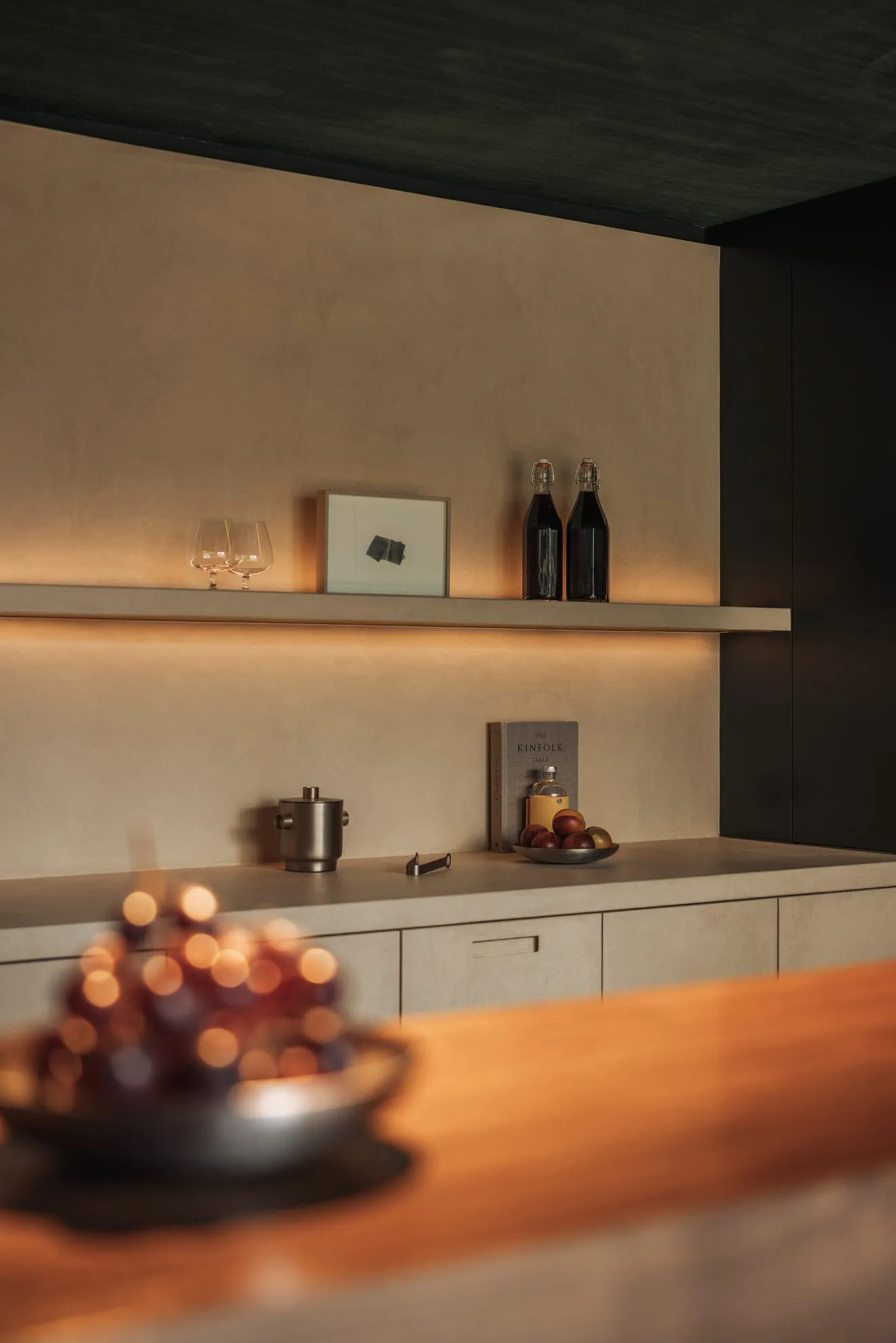A kitchen countertop with wine glasses, bottles, apples, an orange, and a bowl of fruit, creating a cozy and inviting atmosphere