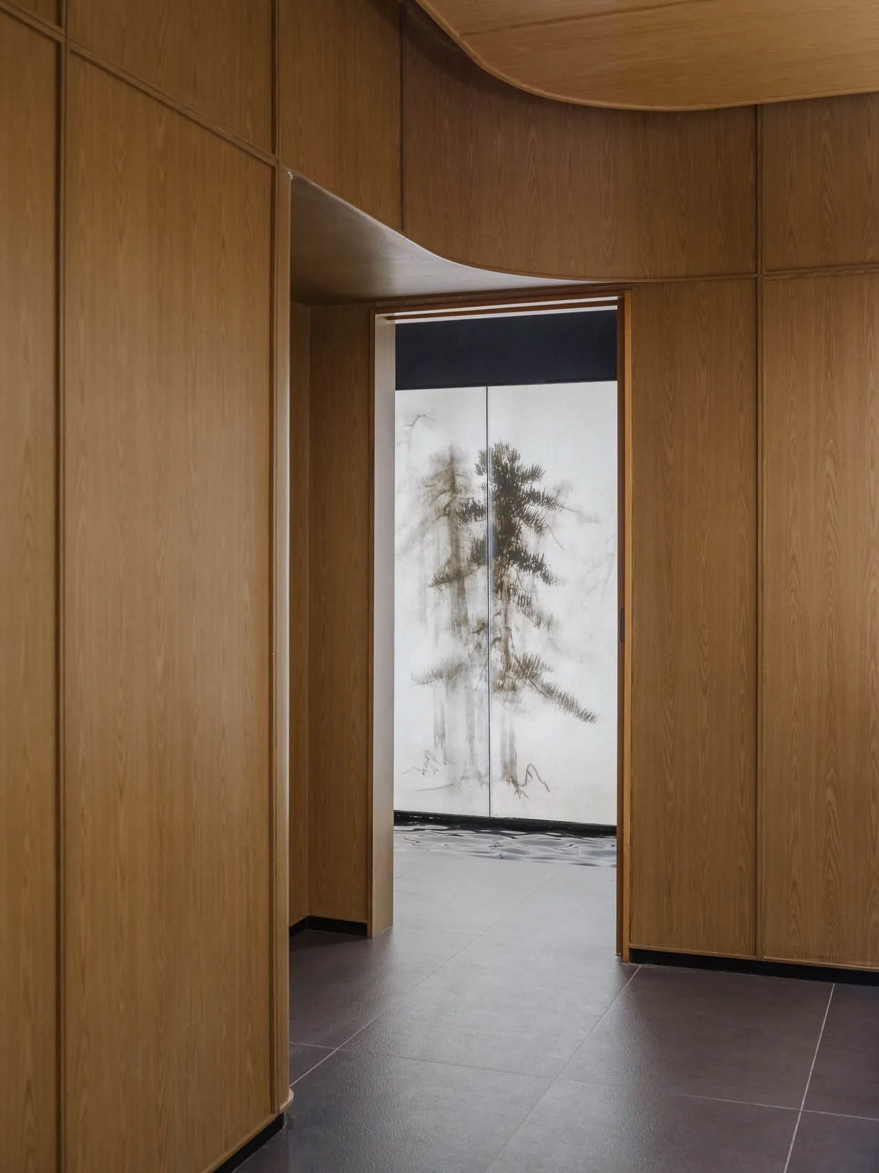 A well-lit interior hallway with wooden walls and floors, featuring a painting of trees on one door and a bright atmosphere