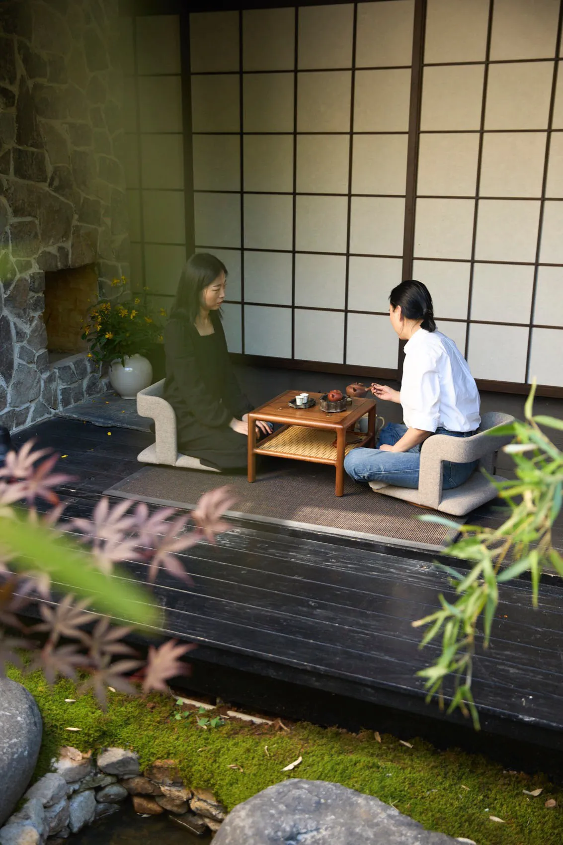 Two women sit in an Asian-style living area with a wooden table, surrounded by decorative vases and potted plants, creating a cozy atmosphere