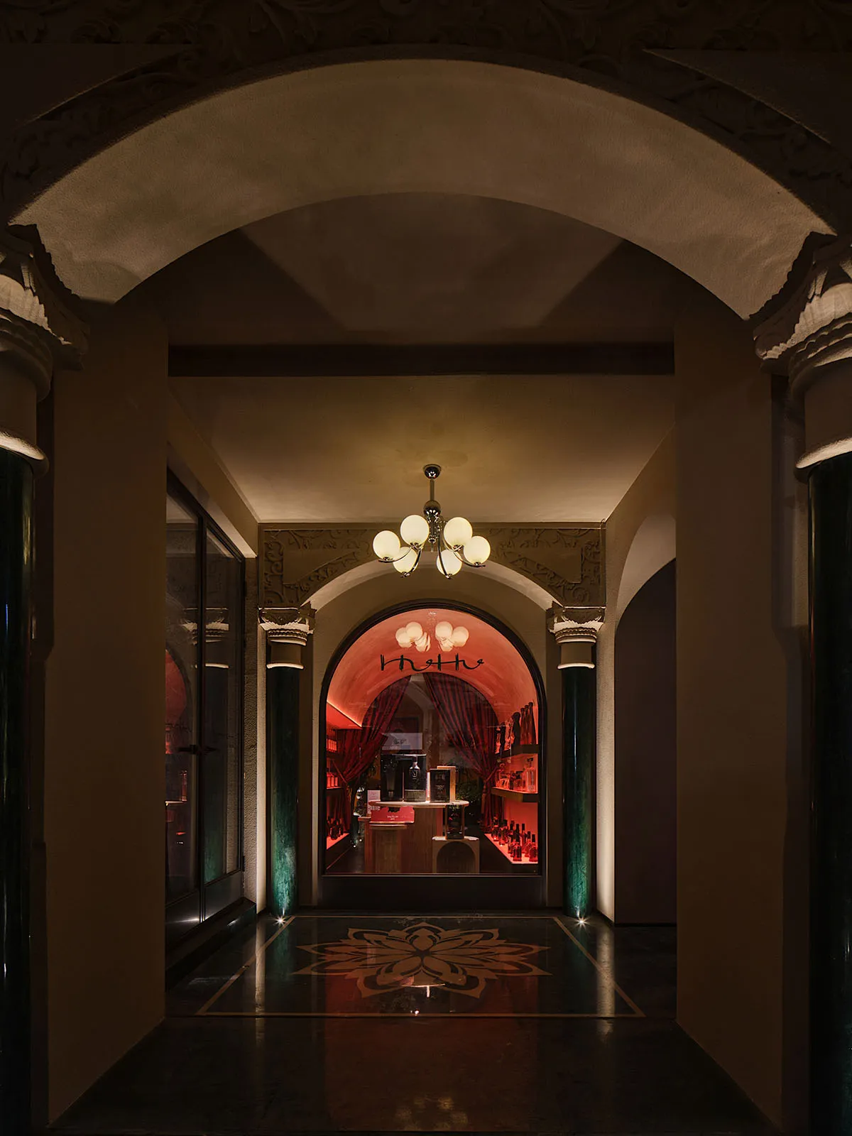 Ornate hallway with red walls, a grand archway, and chandeliers providing illumination