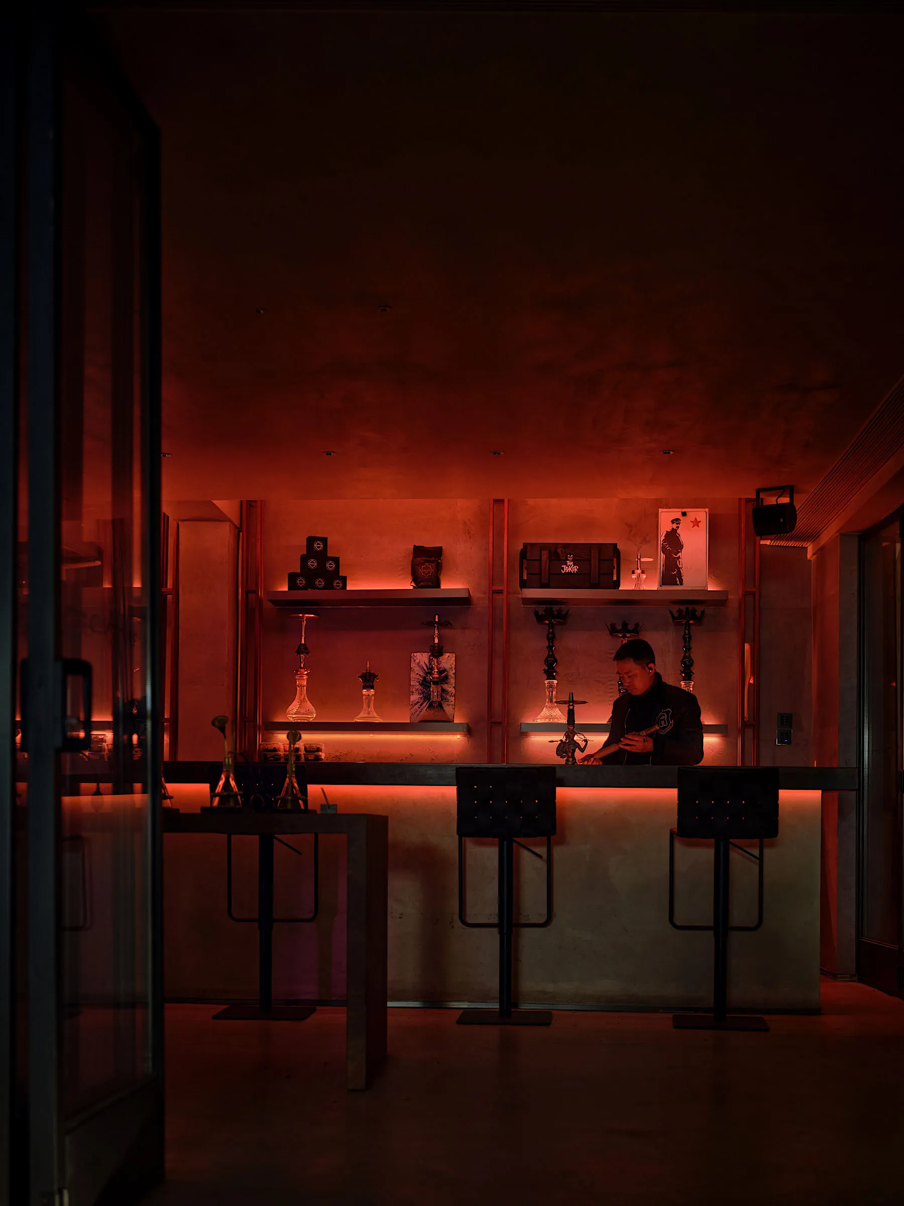 Dimly lit bar area with a stone-like wall, a counter, and stools, featuring bottles and a modern ambiance