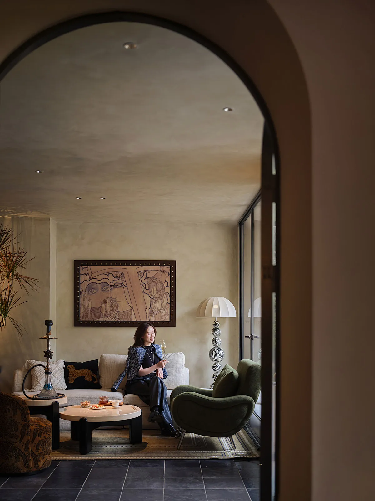 A woman sits in a cozy living room with a dining table, couches, a potted plant, and a vase, surrounded by warm and inviting decor