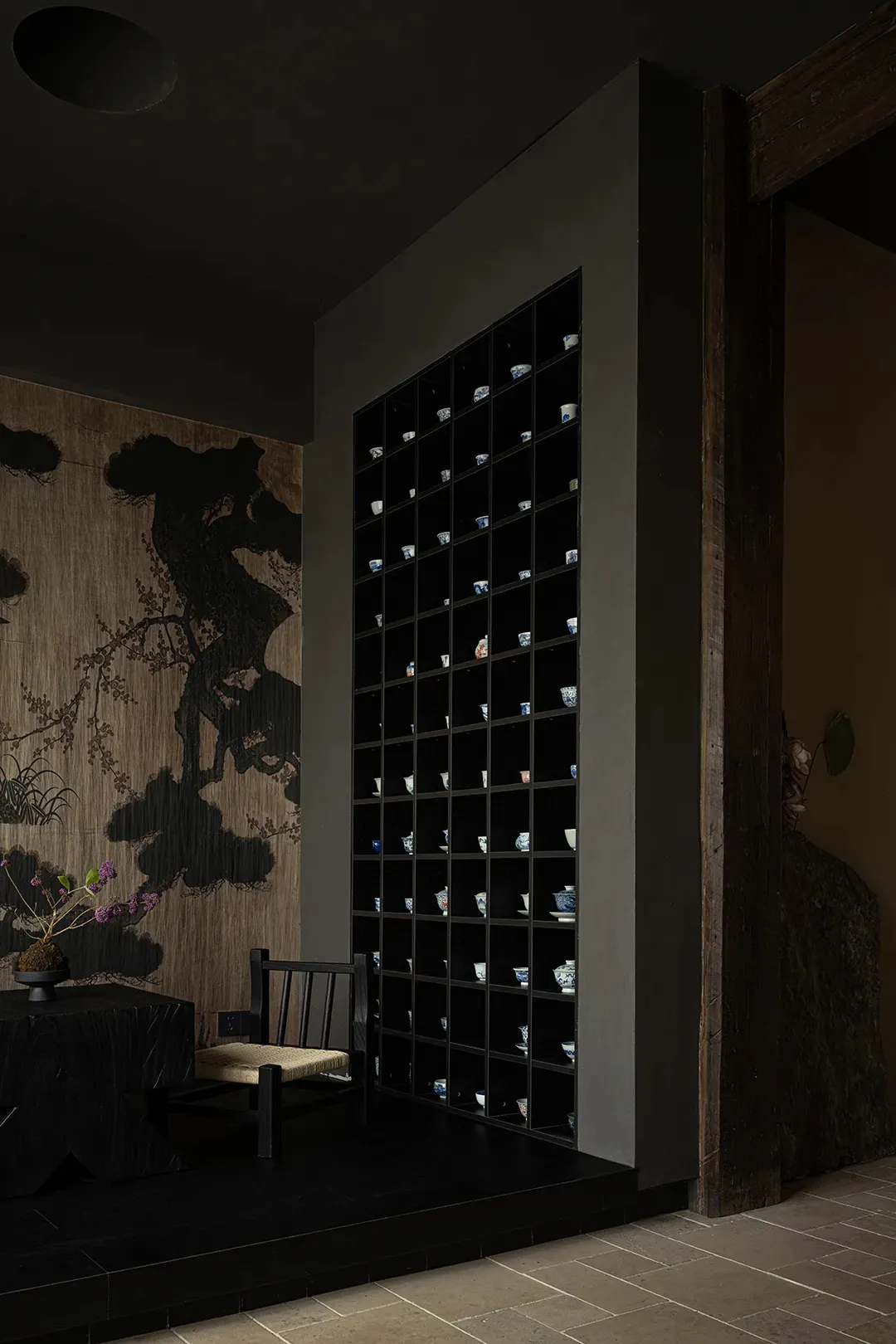 A darkly lit room with Asian-inspired decor, featuring vases on tables and walls, and scattered cups and bowls, with a chair in the corner