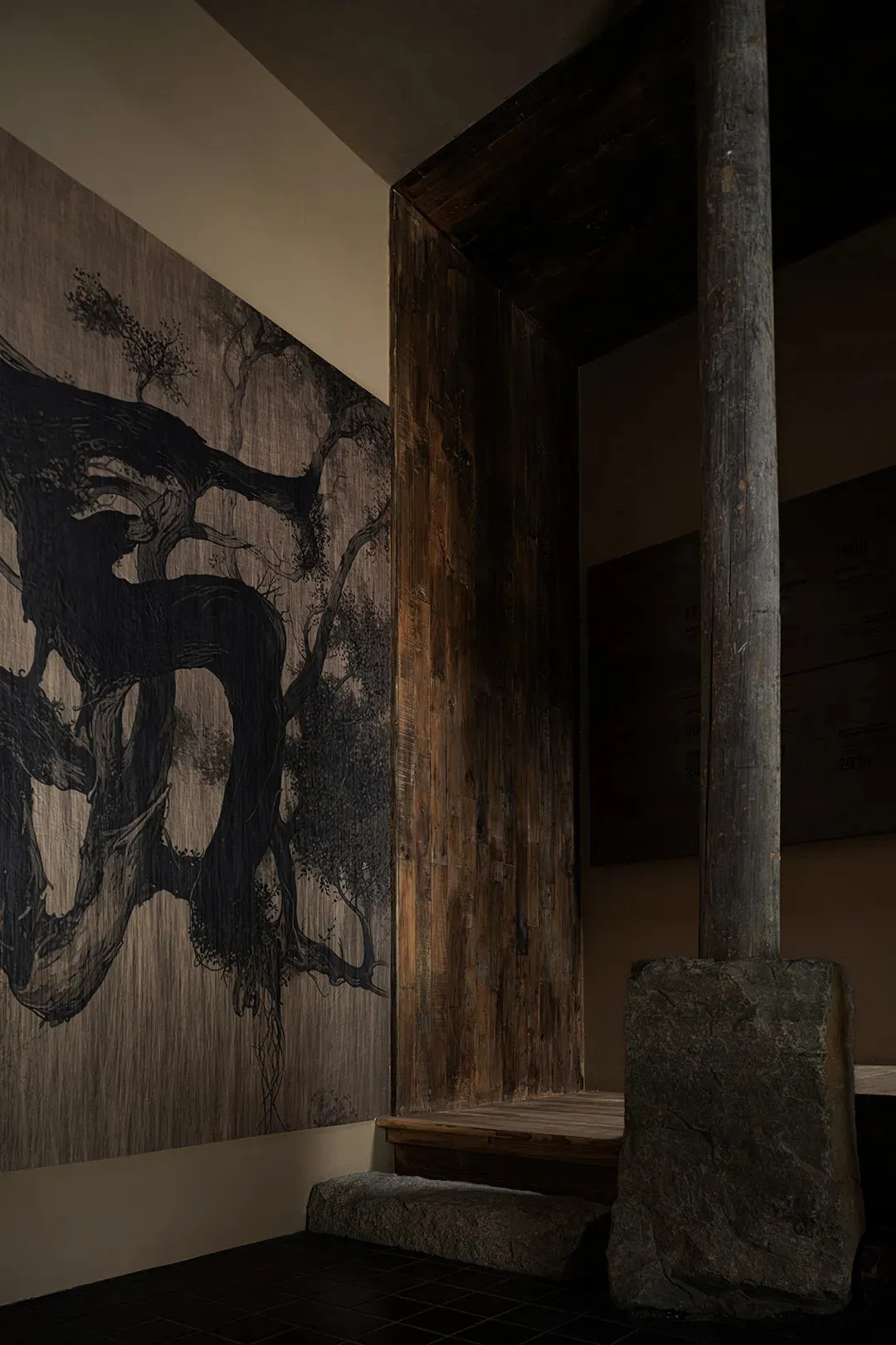 A darkened room features an abstract painting of a tree on the wall, created with wood and paint, in predominantly black and brown hues, with a moody atmosphere