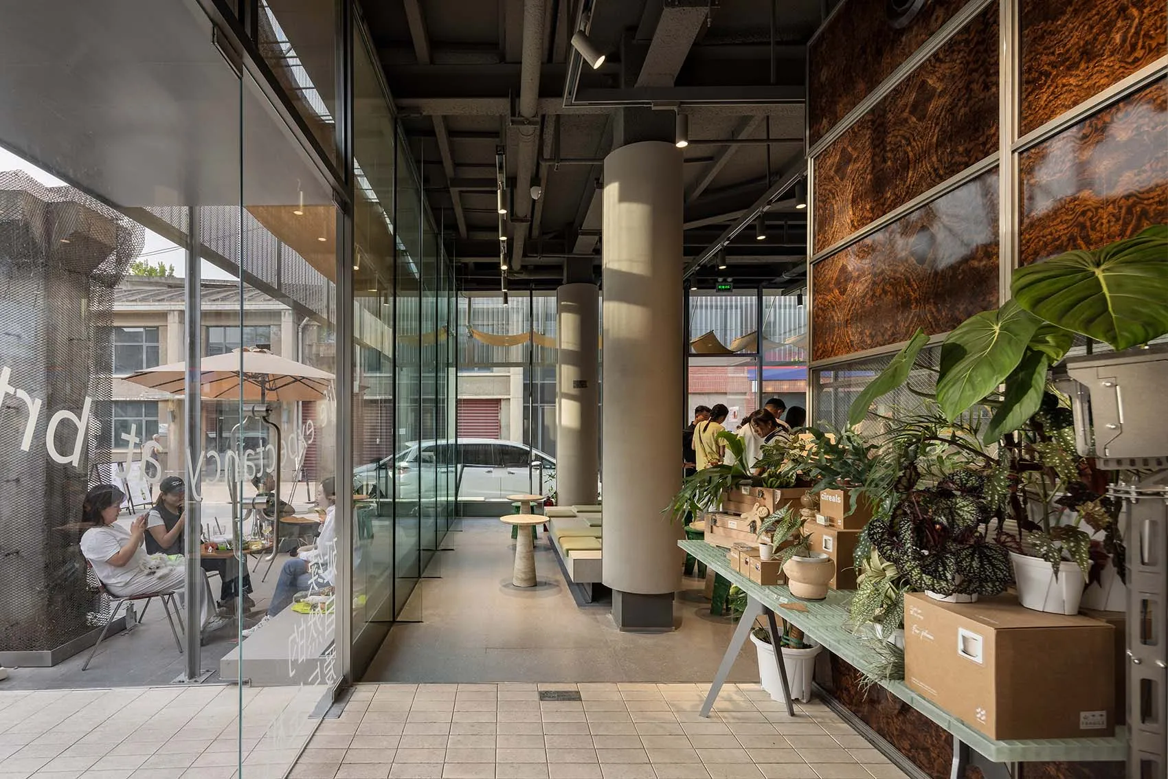 Bright and inviting cafe with glass walls, wooden furniture, and potted plants, featuring multiple dining tables and people socializing