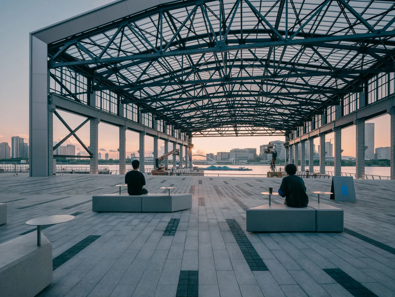 Two men sitting on benches in a modern industrial cafe with a metal roof and a waterfront view of several boats