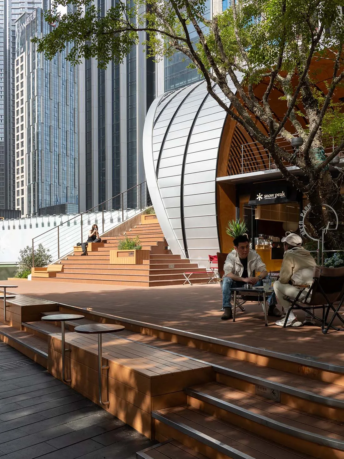 大型金屬牆面建築前方休閒戶外場景，人們坐在椅子上與長椅上，附近停放自行車