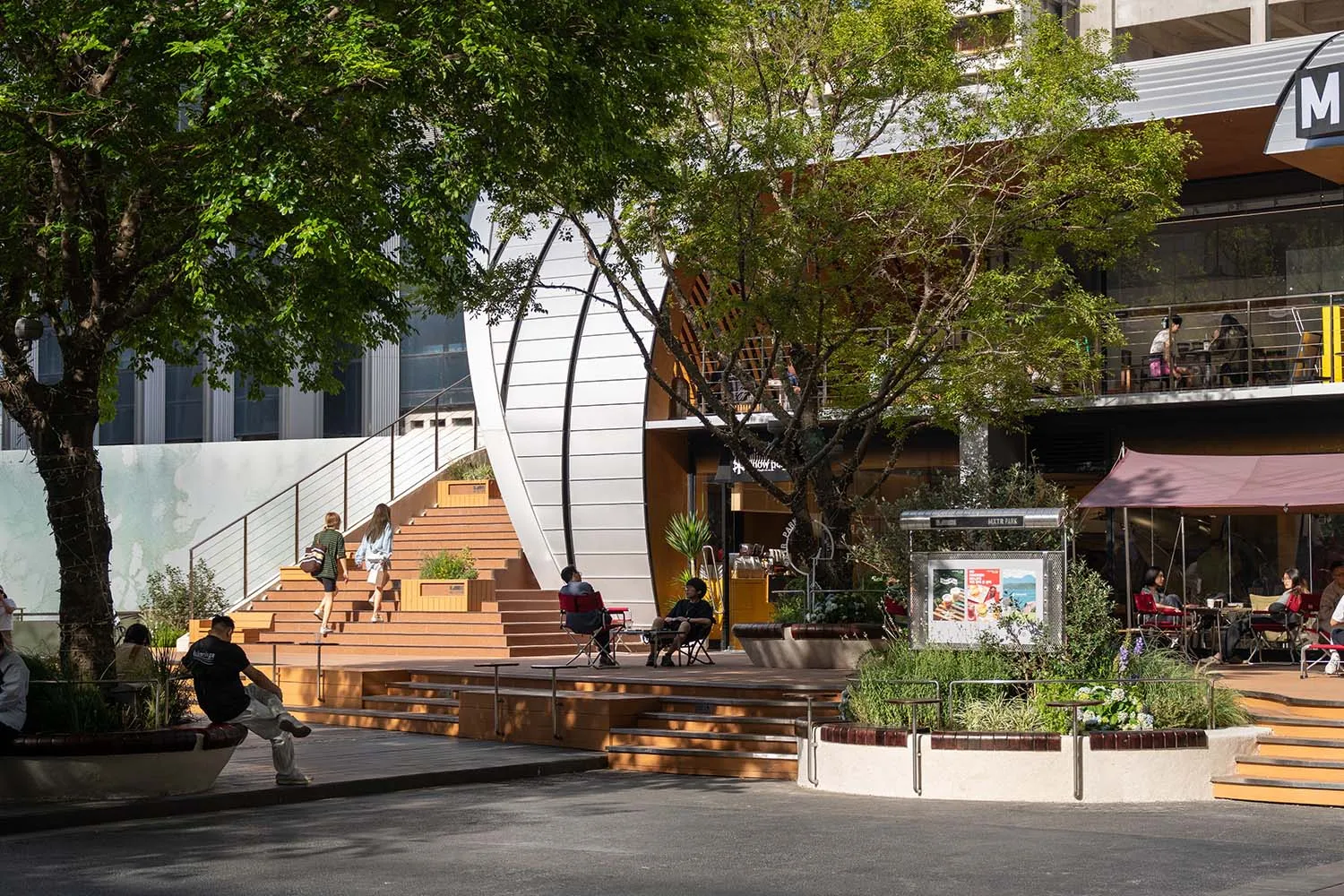 A bustling urban park scene with a large circular structure surrounded by trees, featuring pedestrians, chairs, and lively atmosphere