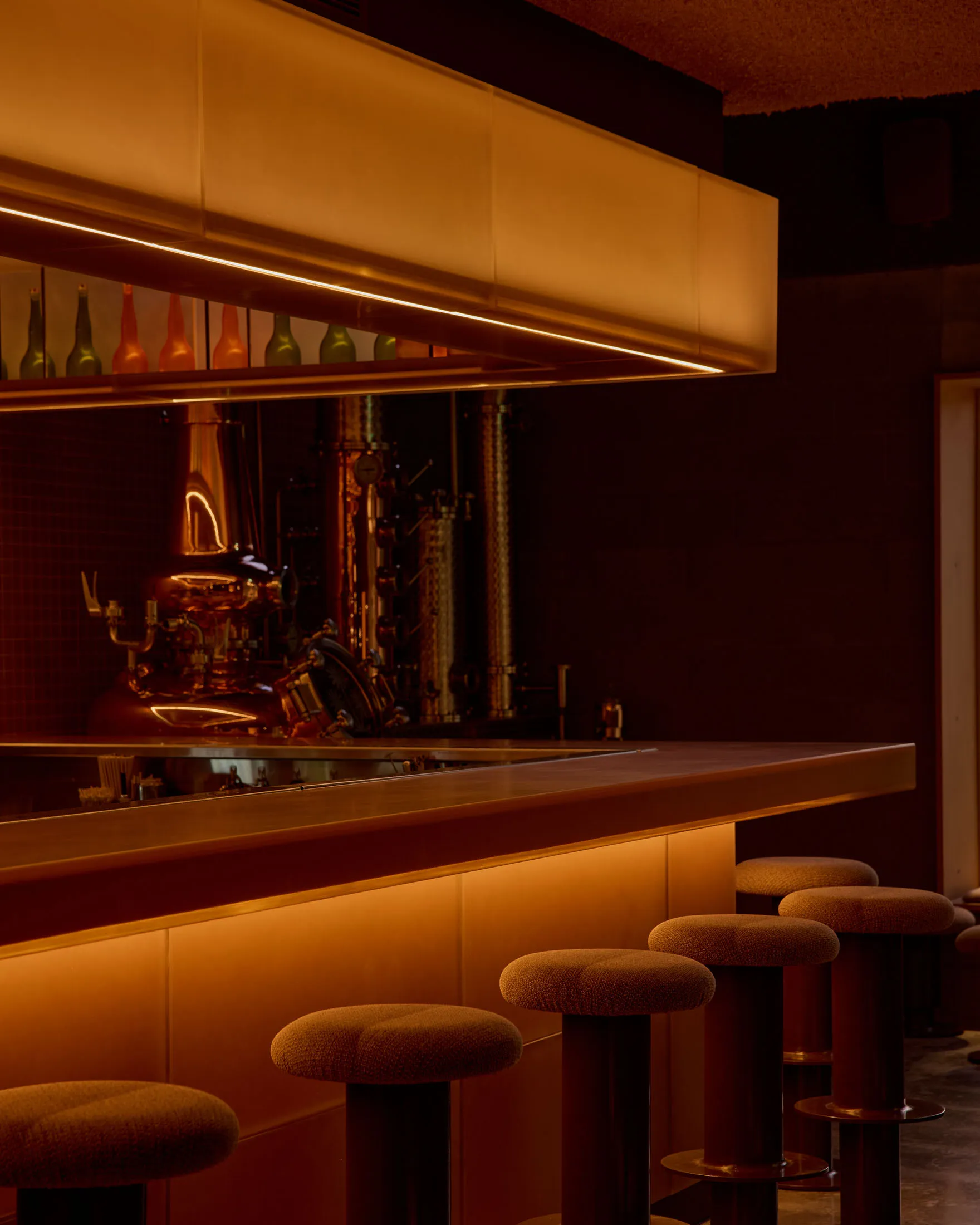 A dimly lit bar with a modern design, featuring a large counter with stools and bottles on display, in a warm atmosphere with brown and orange tones
