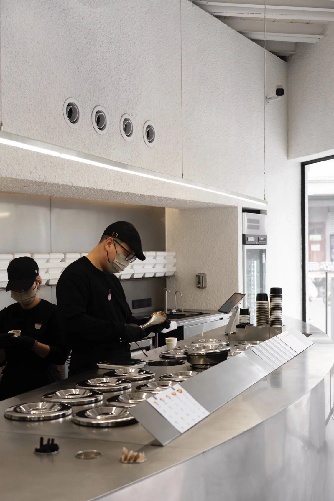 Industrial kitchen with two people working, wearing face masks, surrounded by appliances, bowls, and cups