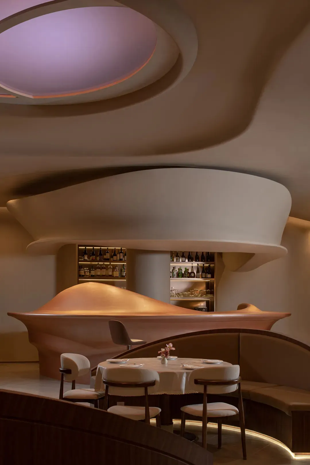 Modern restaurant interior with a curved wall, a dining table, and chairs, featuring bottles on the countertop