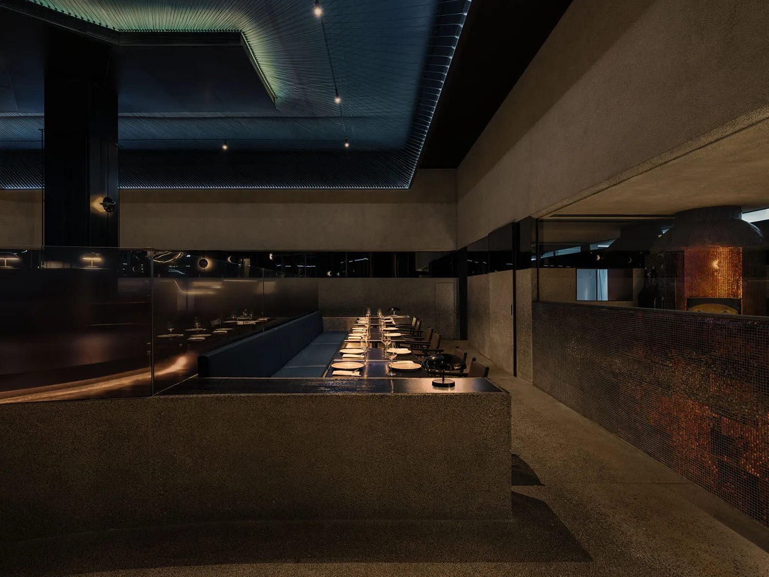 Dimly lit industrial kitchen with a long concrete countertop and multiple chairs, featuring several bowls and a modern aesthetic