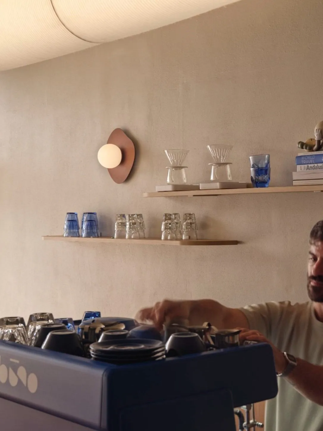 A man stands in a kitchen surrounded by cups, bowls, bottles, and books on various surfaces, creating a lived-in environment