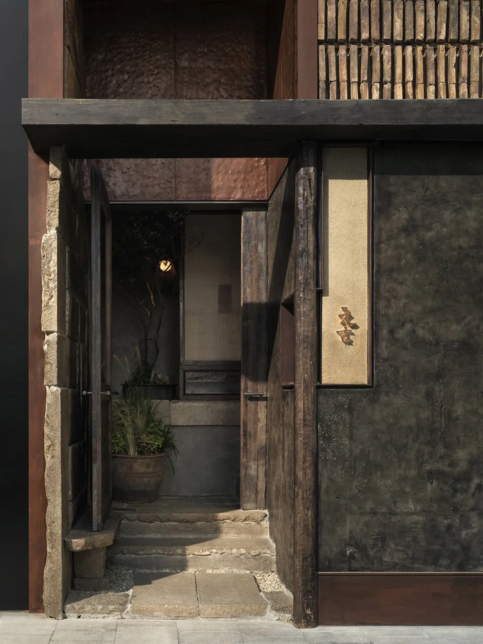 A traditional Asian house with a wooden doorway leading to a small courtyard, featuring stone walls, black walls, and a potted plant near the entrance