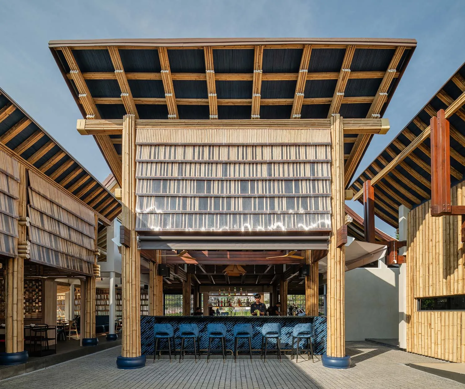 An open-air restaurant with a wooden roof, a bar area, and multiple chairs, surrounded by people enjoying a relaxed atmosphere
